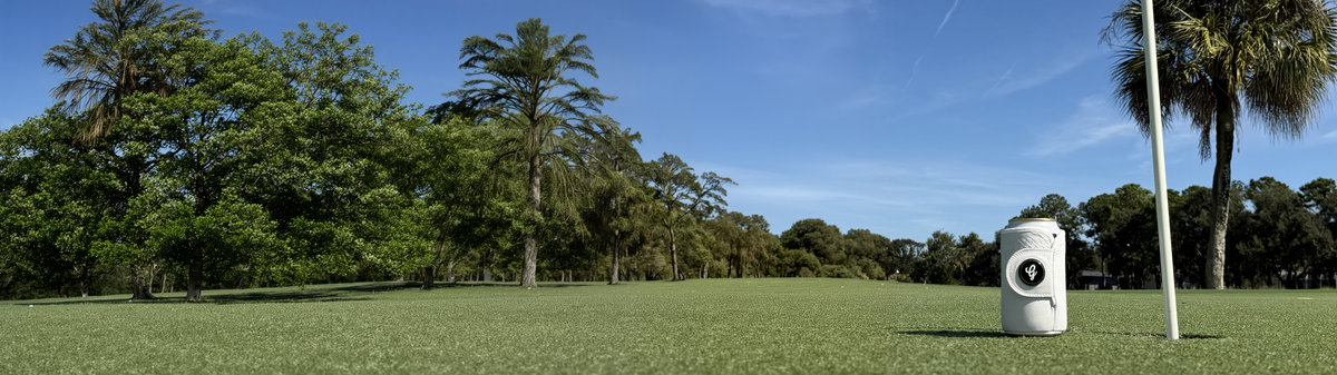 White Gloozie golf glove coozie beside a golf hole on a sunny manicured golf course with palm trees and blue sky