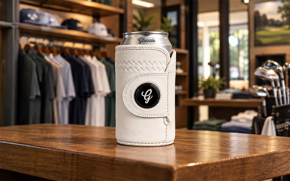 White Gloozie golf glove coozie displayed on a wooden counter in a golf apparel shop with golf clubs and clothing in the background